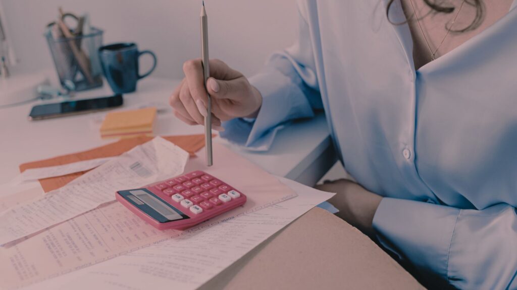 A desk with a calculator and bills with a person holding a pencil above it.